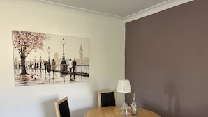 A dining area with a round wooden table, two chairs, a lamp, and a painting of a cityscape.