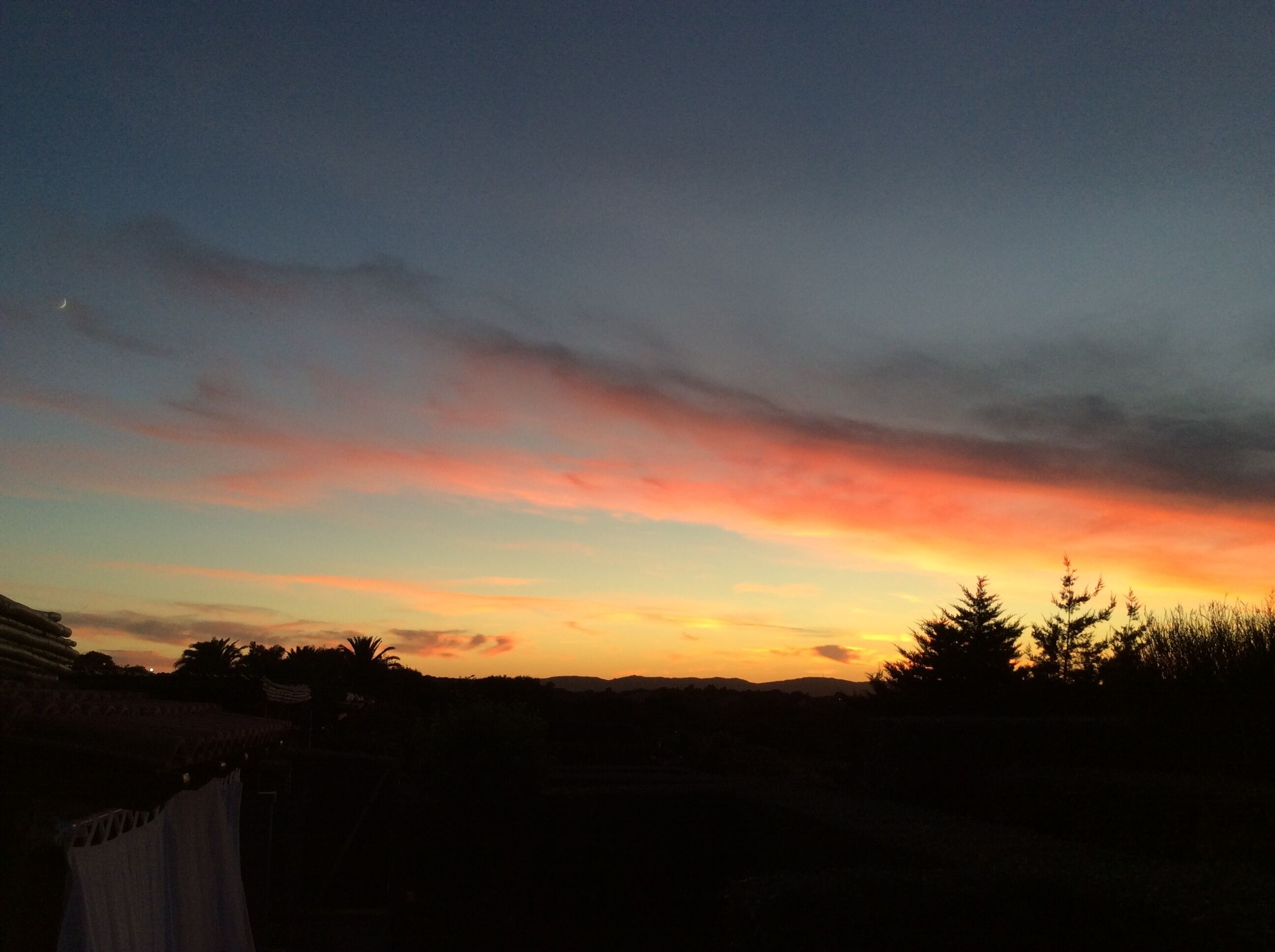 Un coucher de soleil avec un ciel dégagé et les silhouettes d’arbres et d’un bâtiment.