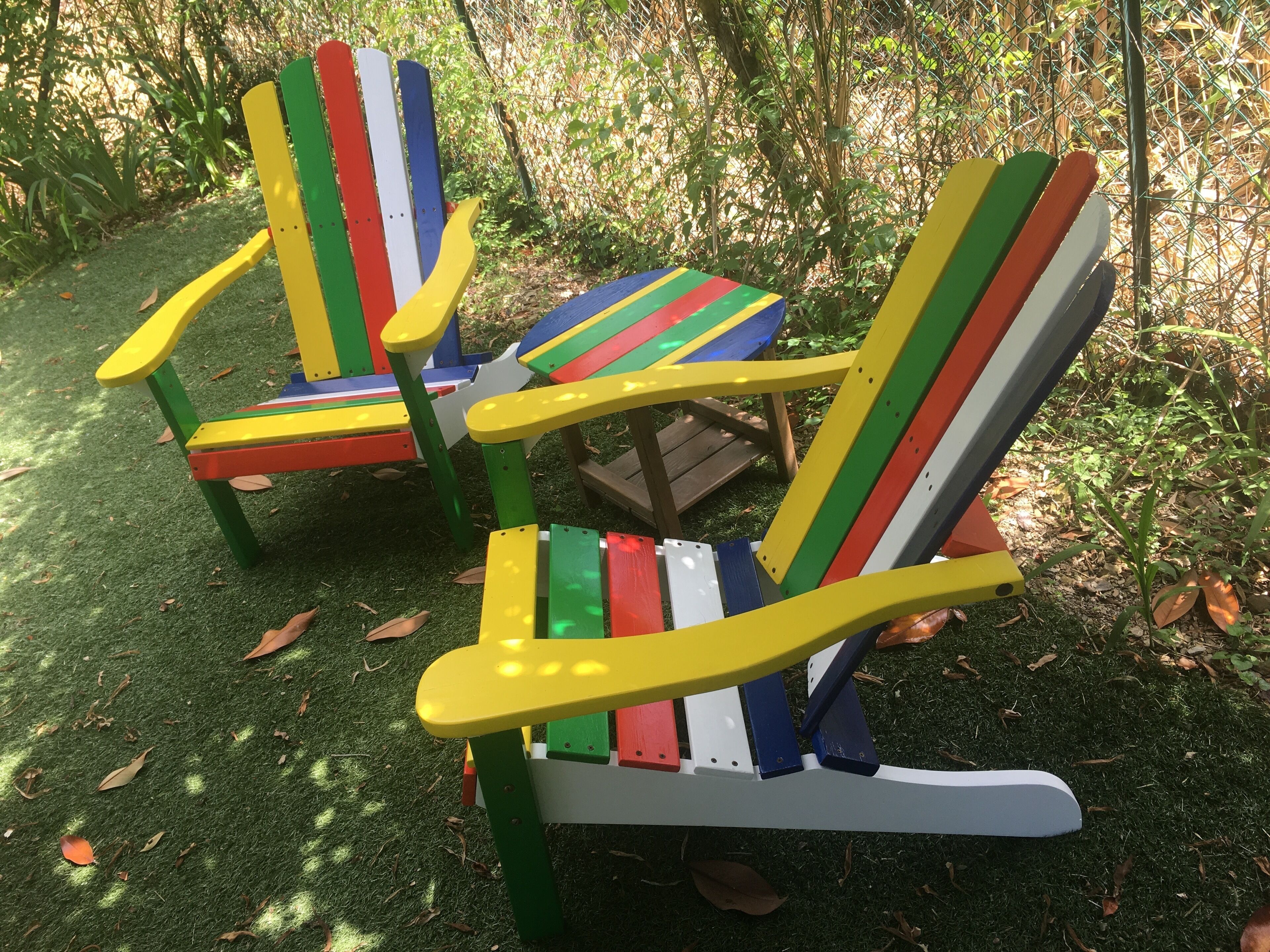 Two colorful Adirondack chairs with a small table in between, set on a grassy area with trees in the background.