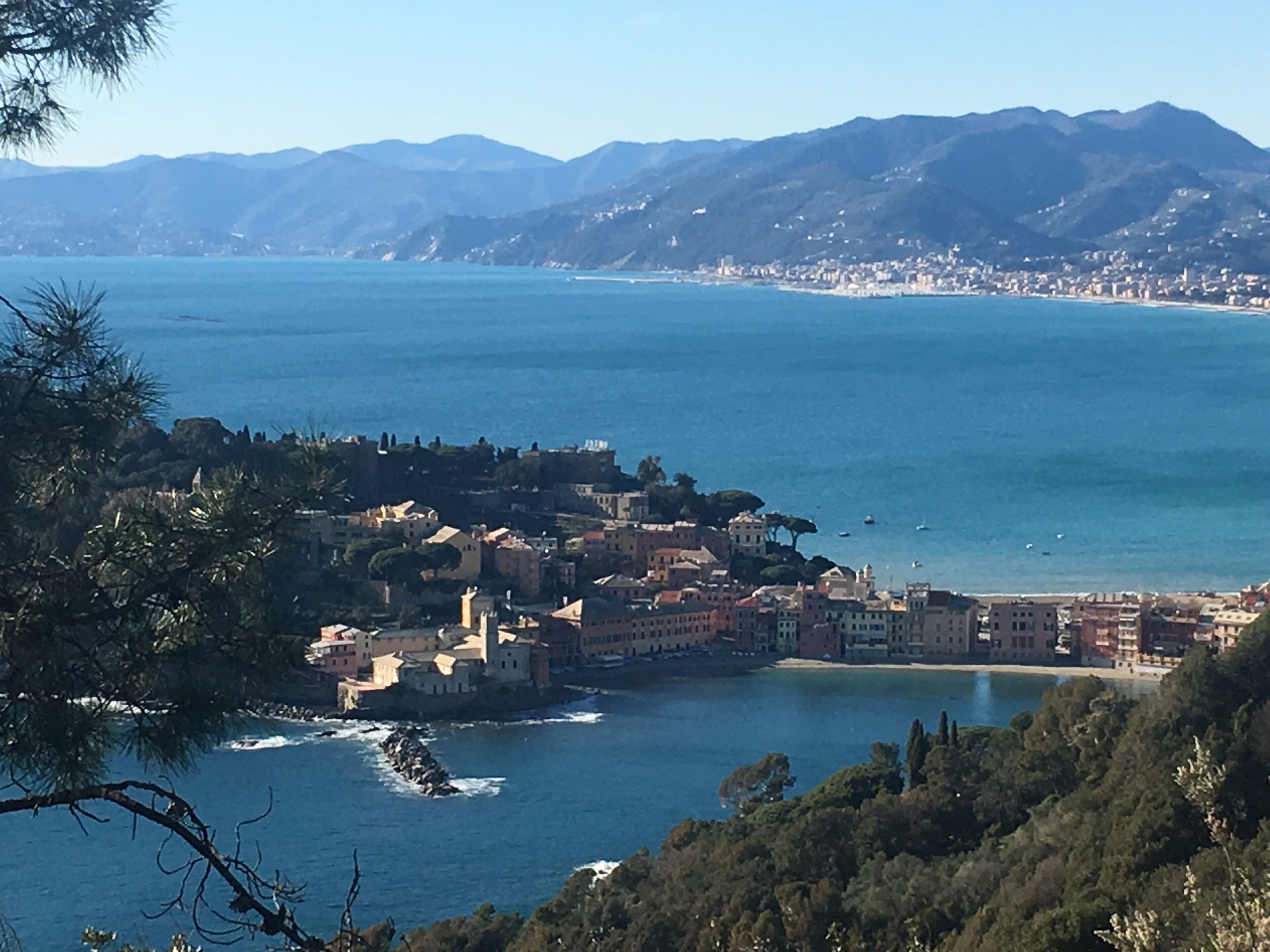 A coastal town with buildings along the shoreline, a clear view of the sea, and mountains in the background.