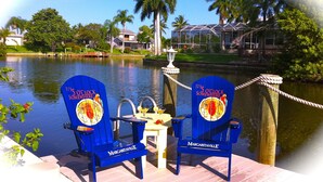Two blue Adirondack chairs with 'Margaritaville' branding on a dock by a lake, surrounded by palm trees and houses. - VILLA SUNSET KISS•SW•DREAMLESS BEAUTIFUL•CHANNEL CROSSING WITH SEA ACCESS ***** (Cape Coral)