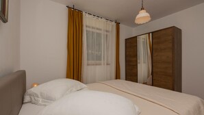 Et soveværelse med en seng, to puder, et træskab, et vindue med let gardin og en hængende lampe.