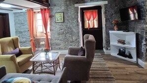 A rustic interior with exposed wooden beams, stone walls, a sofa, armchairs, a coffee table, and a television on a shelf. - Rural Tourism - Laurissilva Forest (Camacha)