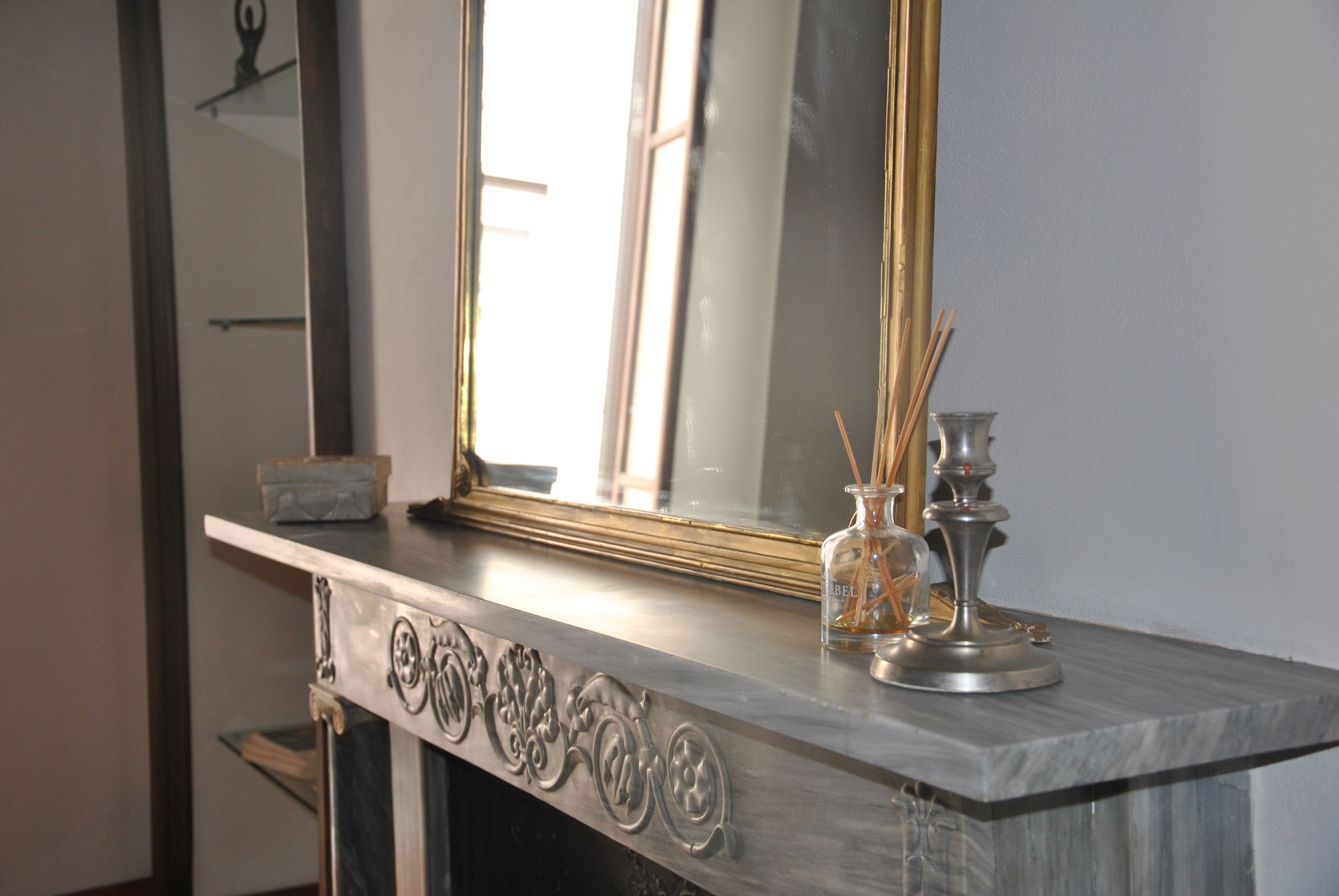 A marble fireplace with a large mirror above it, a small vase with reed sticks, and a candle holder.