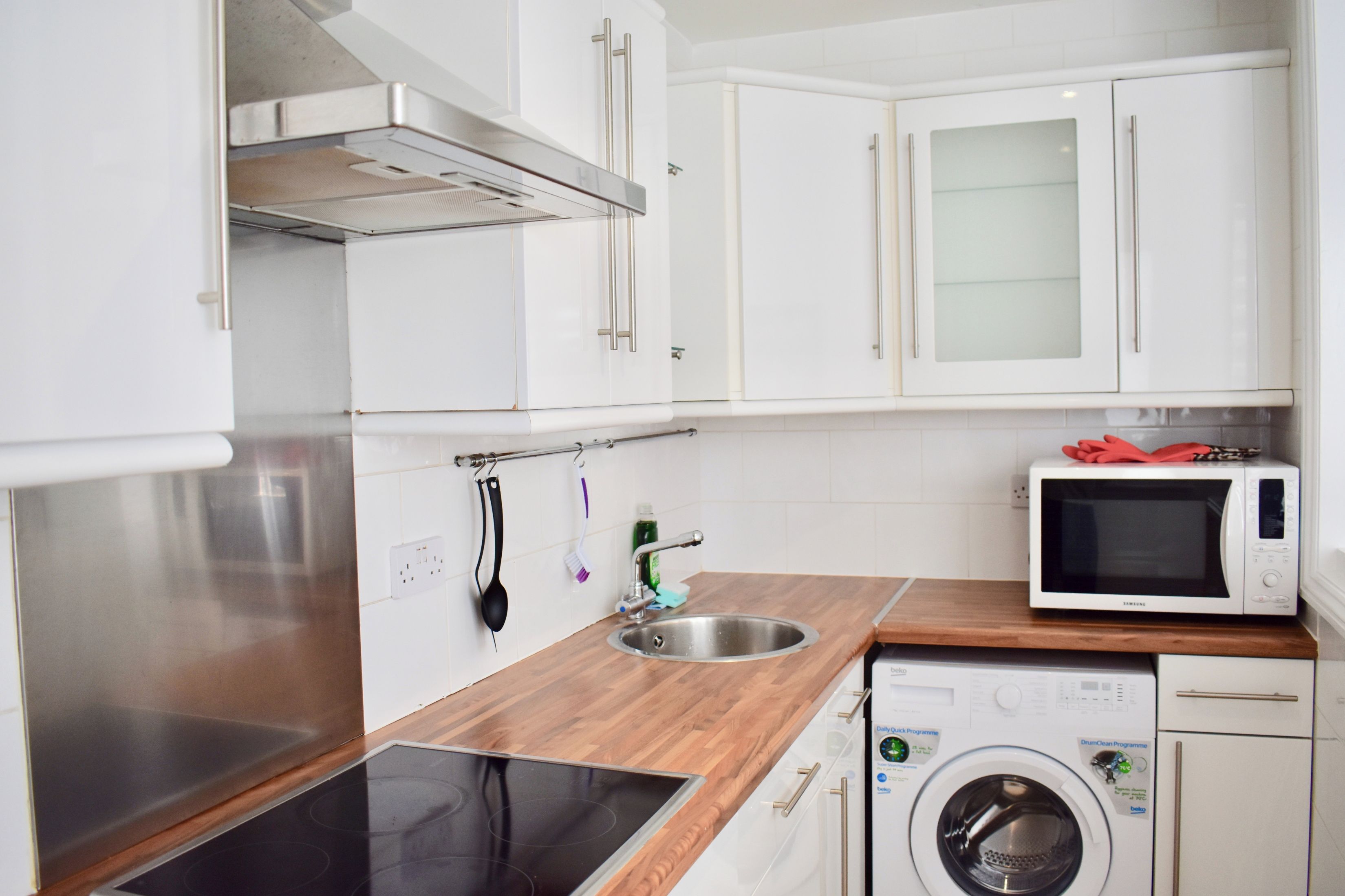 A modern kitchen with white cabinets, a stainless steel sink, a microwave, a built-in oven, and a washing machine.