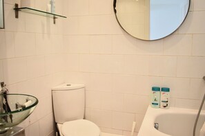 Un baño con inodoro, lavabo de cristal, espejo redondo y dos botellas de champú en una repisa.