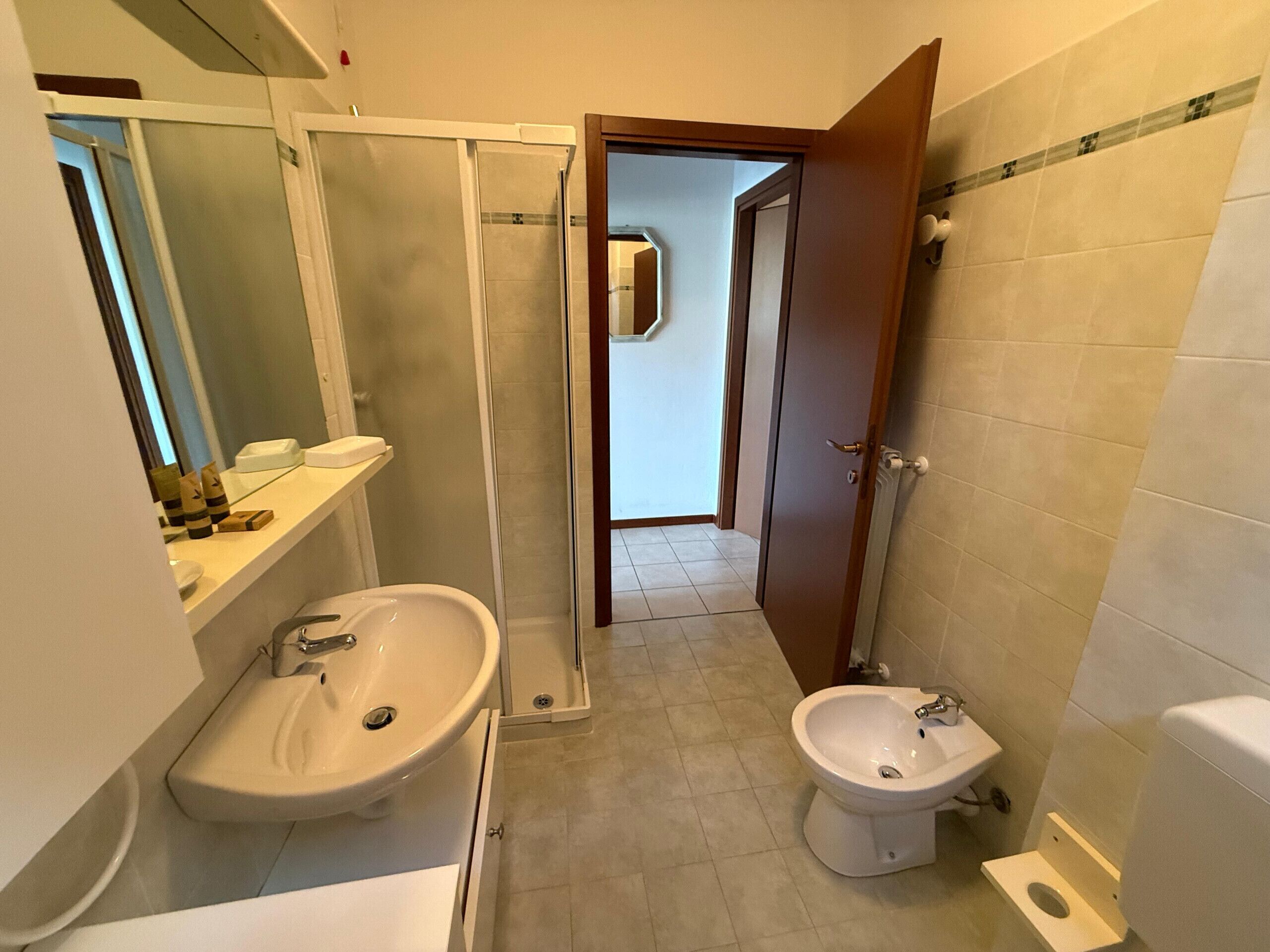Um banheiro pequeno com pia, bidê e vaso sanitário. Há um espelho sobre a pia e uma porta de chuveiro que dá para um corredor.