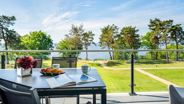 Una mesa de comedor con un cuenco de fruta, un jarrón de flores y un libro, con vistas a un césped cuidado y una masa de agua.