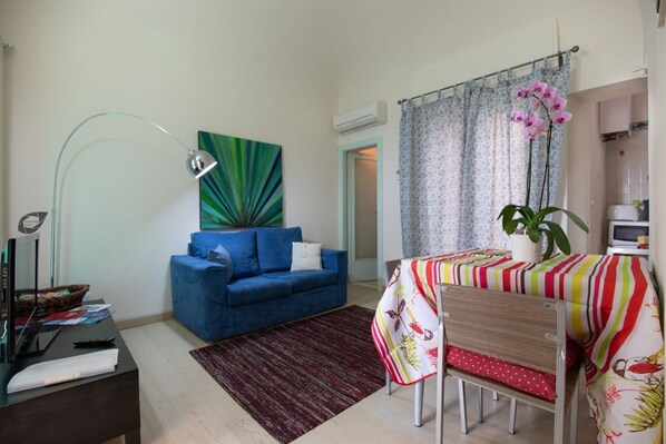 A living room with a blue sofa, a dining table with a colorful tablecloth, a modern floor lamp, and a potted orchid.