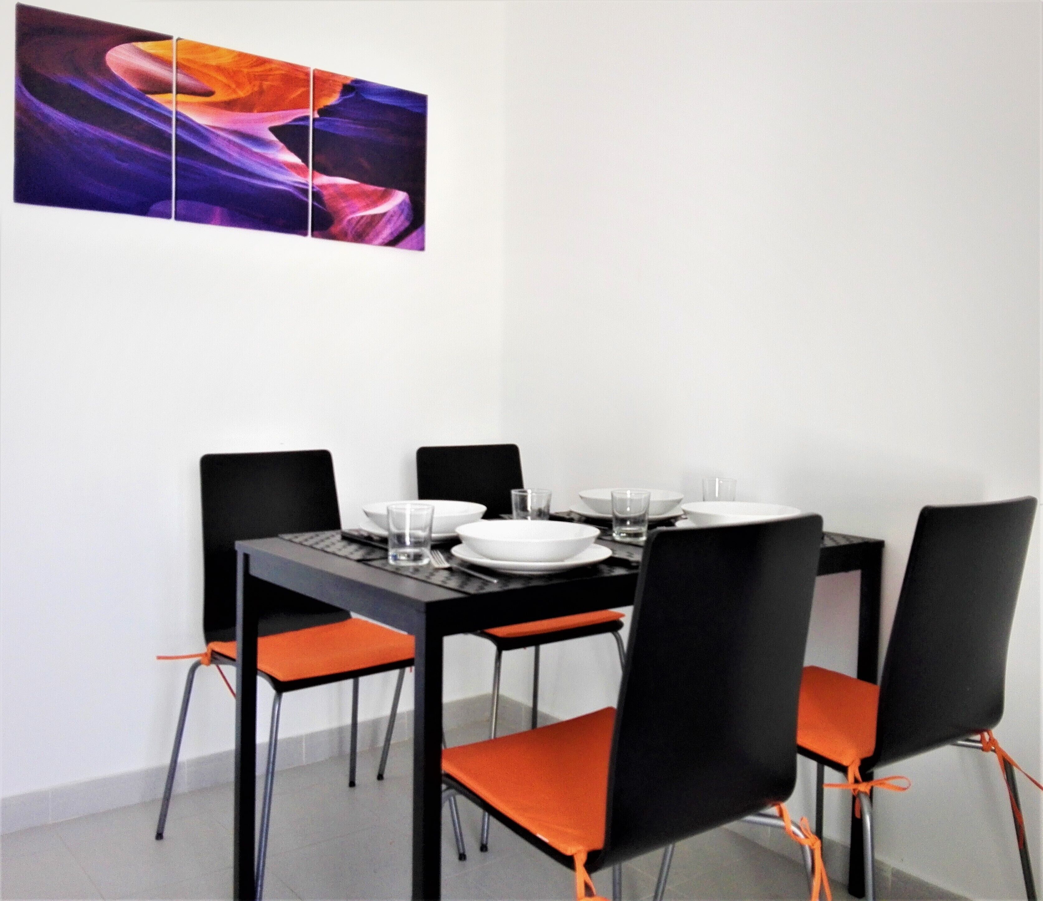 Un comedor con una mesa negra, cuatro sillas negras con cojines anaranjados, platos blancos y vasos transparentes.