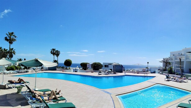 A pool area with lounge chairs and umbrellas, a clear view of the ocean, and a white building with balconies.