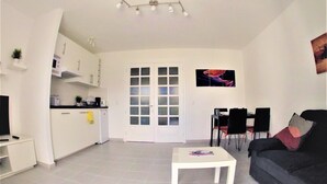 A modern living room with a white coffee table, a dark sofa, and a dining area with chairs. There is a kitchen with white cabinets and a microwave.