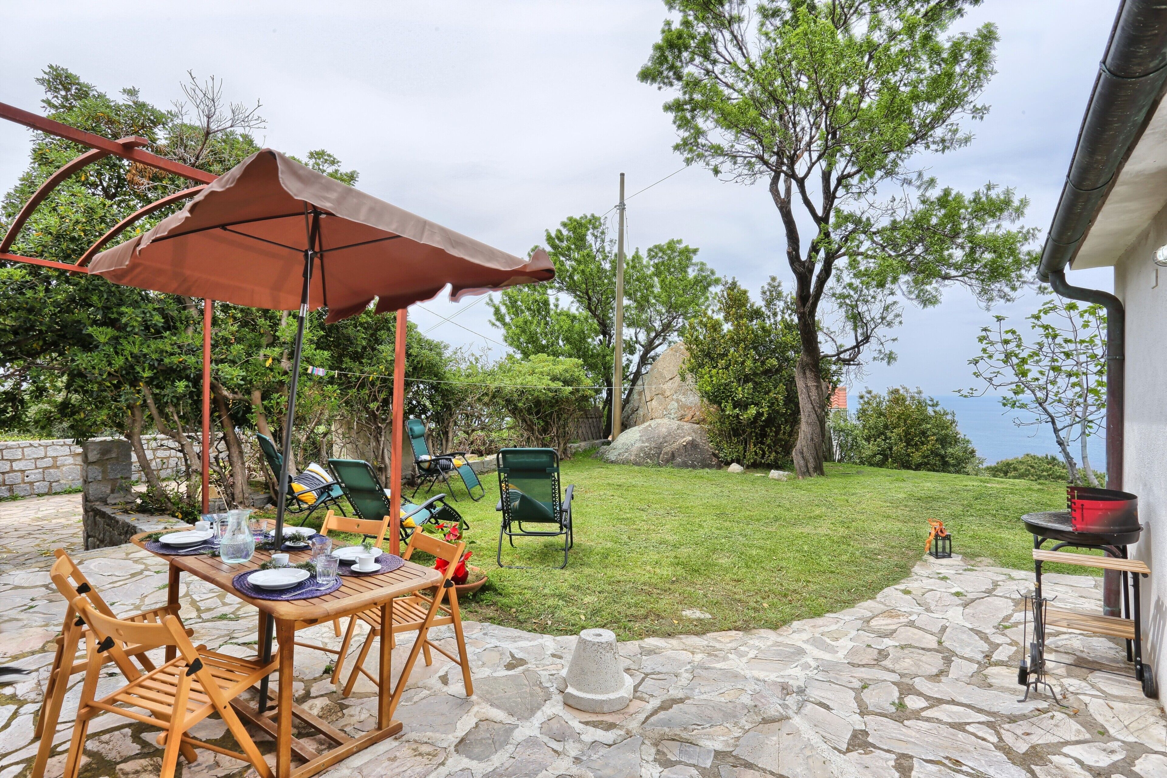 Une terrasse avec une table dressée pour les repas, des chaises d’extérieur, un barbecue et un chemin en pierre.