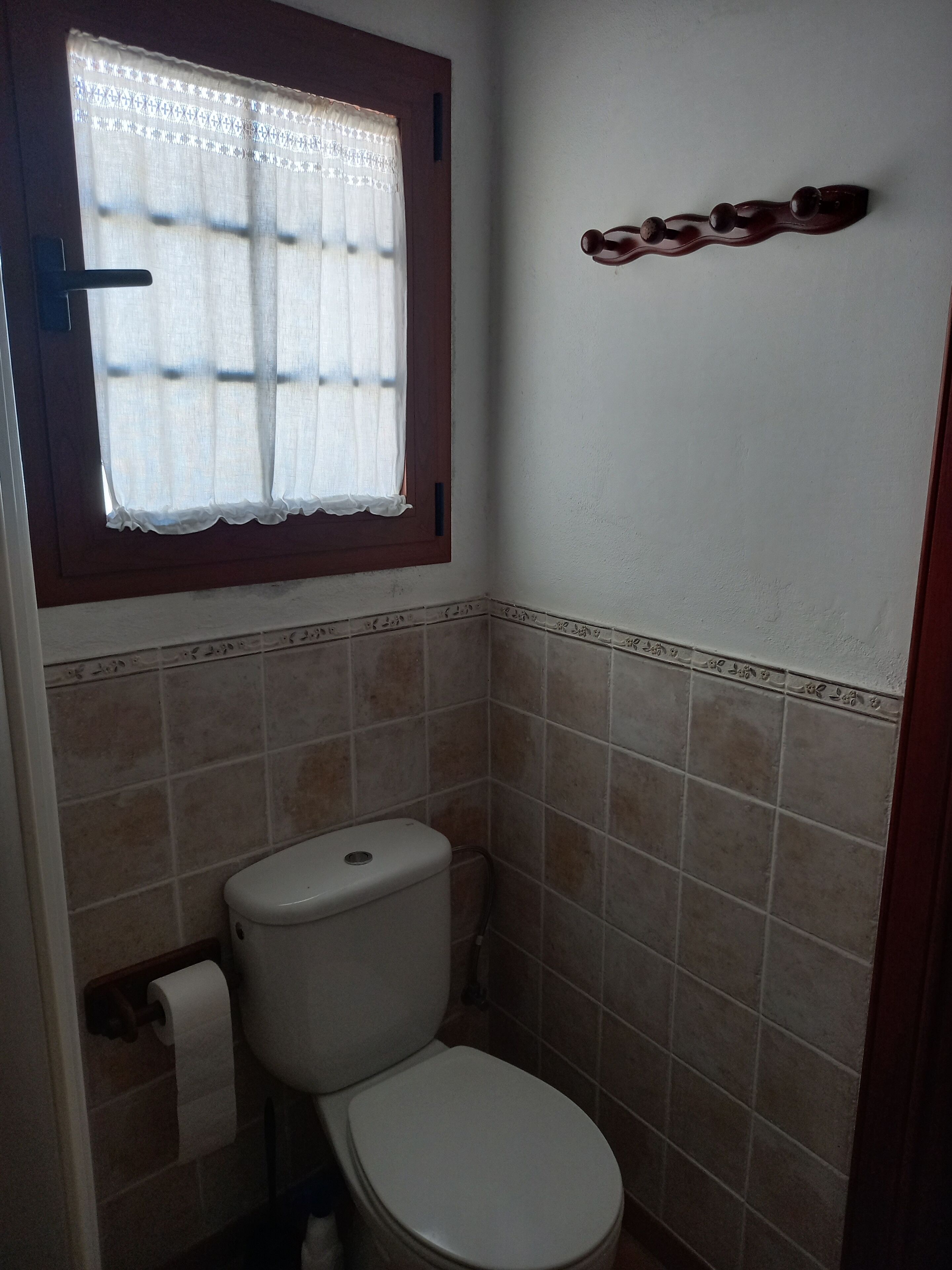 Une petite salle de bain avec des toilettes, une fenêtre ornée d’un rideau en dentelle et un élément décoratif en bois accroché au mur.
