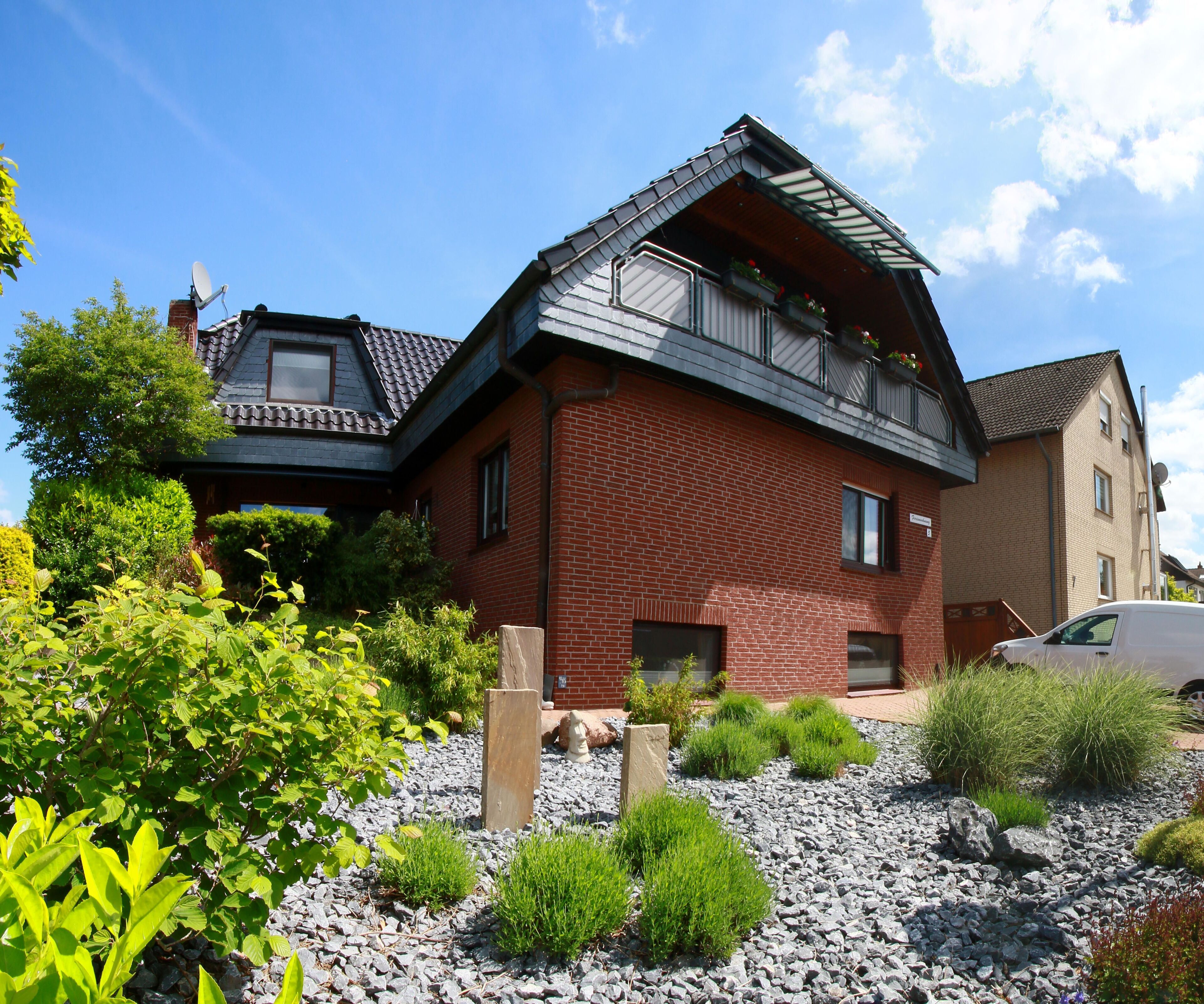 A two-story house with a sloped roof, a balcony with flowers, and a gravel driveway.