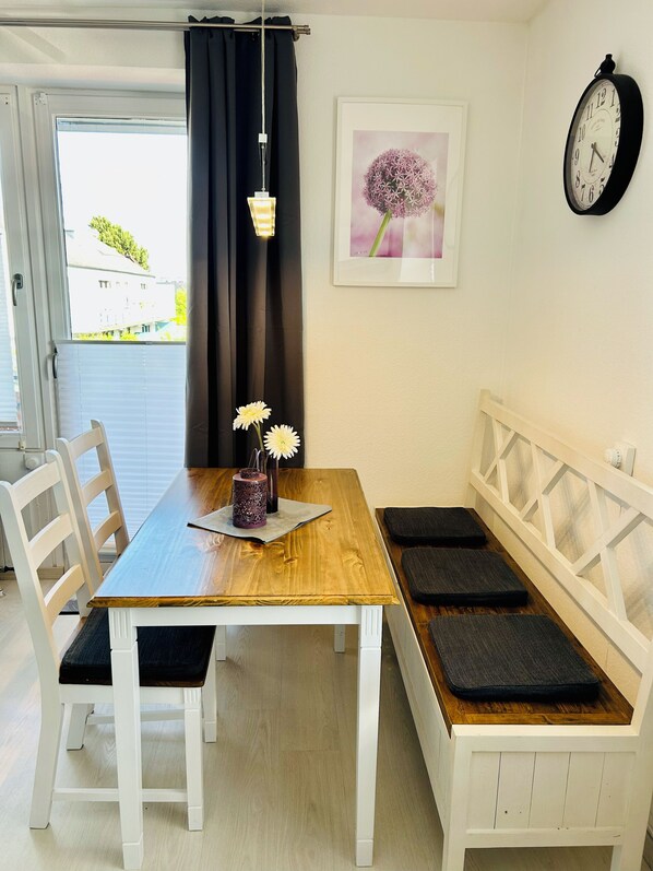 Een eethoek met een houten tafel, witte stoelen, een bank met kussens, een wandklok en een lijst met een afbeelding.