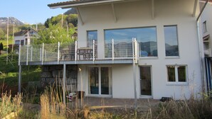 A modern two-story house with a balcony, large windows, and a mountain backdrop.