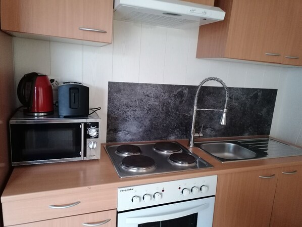 A compact kitchen with a microwave, toaster, kettle, stove, sink, and wooden cabinets.