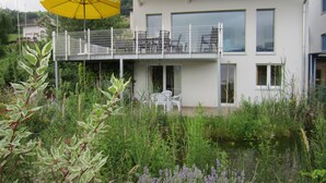 A modern two-story house with a balcony, a yellow umbrella, and a garden with various plants and flowers.