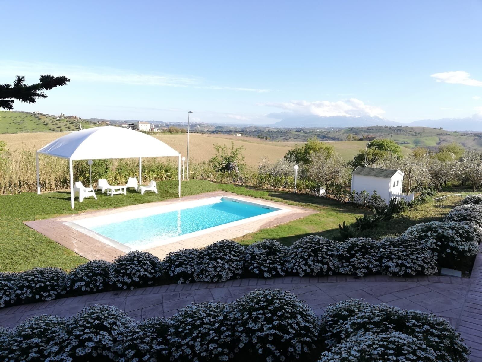 Une piscine surmontée d’un auvent blanc, entourée d’un jardin où fleurissent des plantes aux fleurs blanches, et offrant une vue sur des collines lointaines.