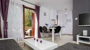 A modern living room with a white sofa, a coffee table, and a dining area with a white table and black chairs. There is a kitchen area with white cabinets and a flat-screen TV mounted on the wall. - 4 star romantic villa, just a minute walk to the beach, heated pool. (Cizici)