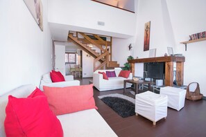 Una sala de estar con una escalera, sofás blancos, cojines rojos, una chimenea y una estantería.