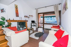 A living room with a fireplace, white sofas with red cushions, a wooden coffee table, and a view of mountains through a large window.