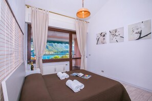 A hotel room with a bed, a window with curtains, a view of a mountain and sea, and artwork on the wall.