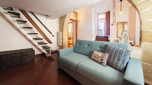 A living room with a blue sofa, a wooden staircase, and a stone wall. - Casa Conchiglia: Authentic Seafront Apartment (Monopoli)