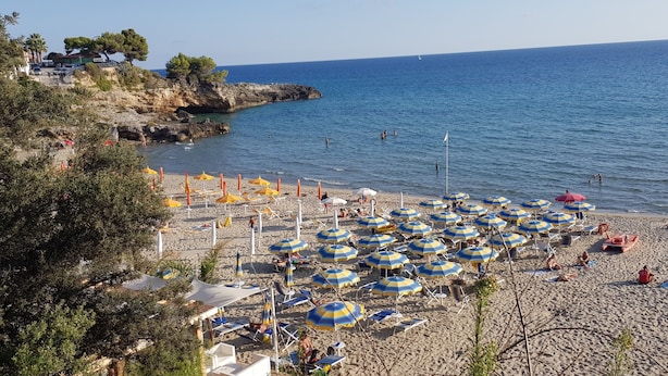 Una playa con numerosas sombrillas y tumbonas, un litoral rocoso y un mar azul cristalino.
