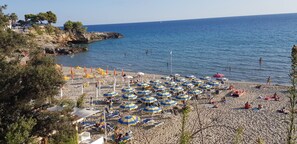 Ein Strand mit vielen Sonnenschirmen und Liegestühlen, einer felsigen Küste und einem klaren, blauen Meer.