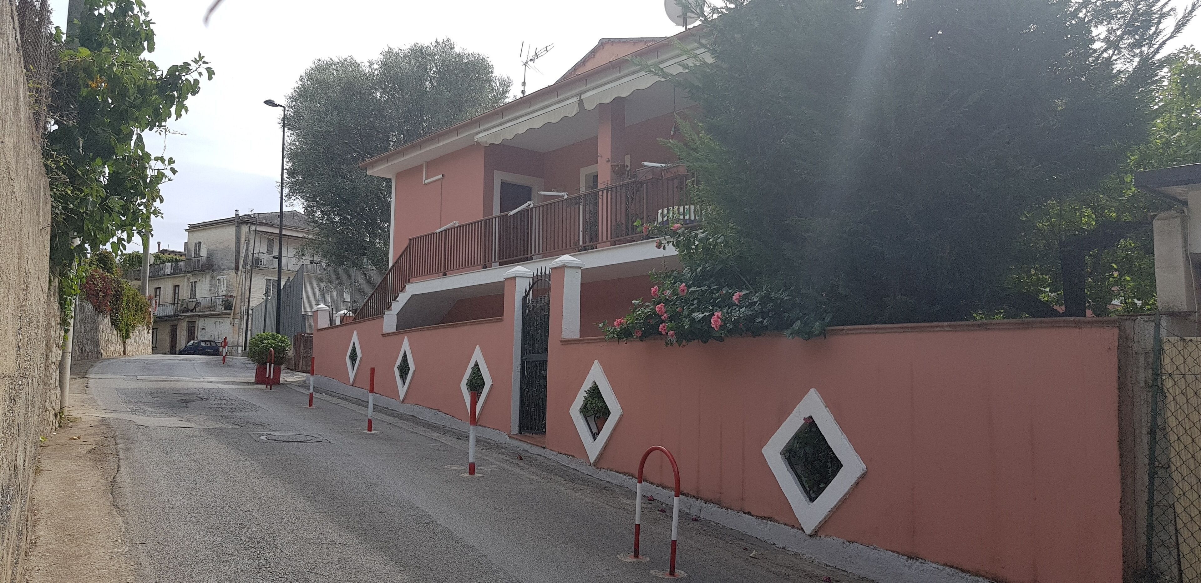 A residential building with a balcony, a red wall with diamond-shaped windows, and a street with bollards.