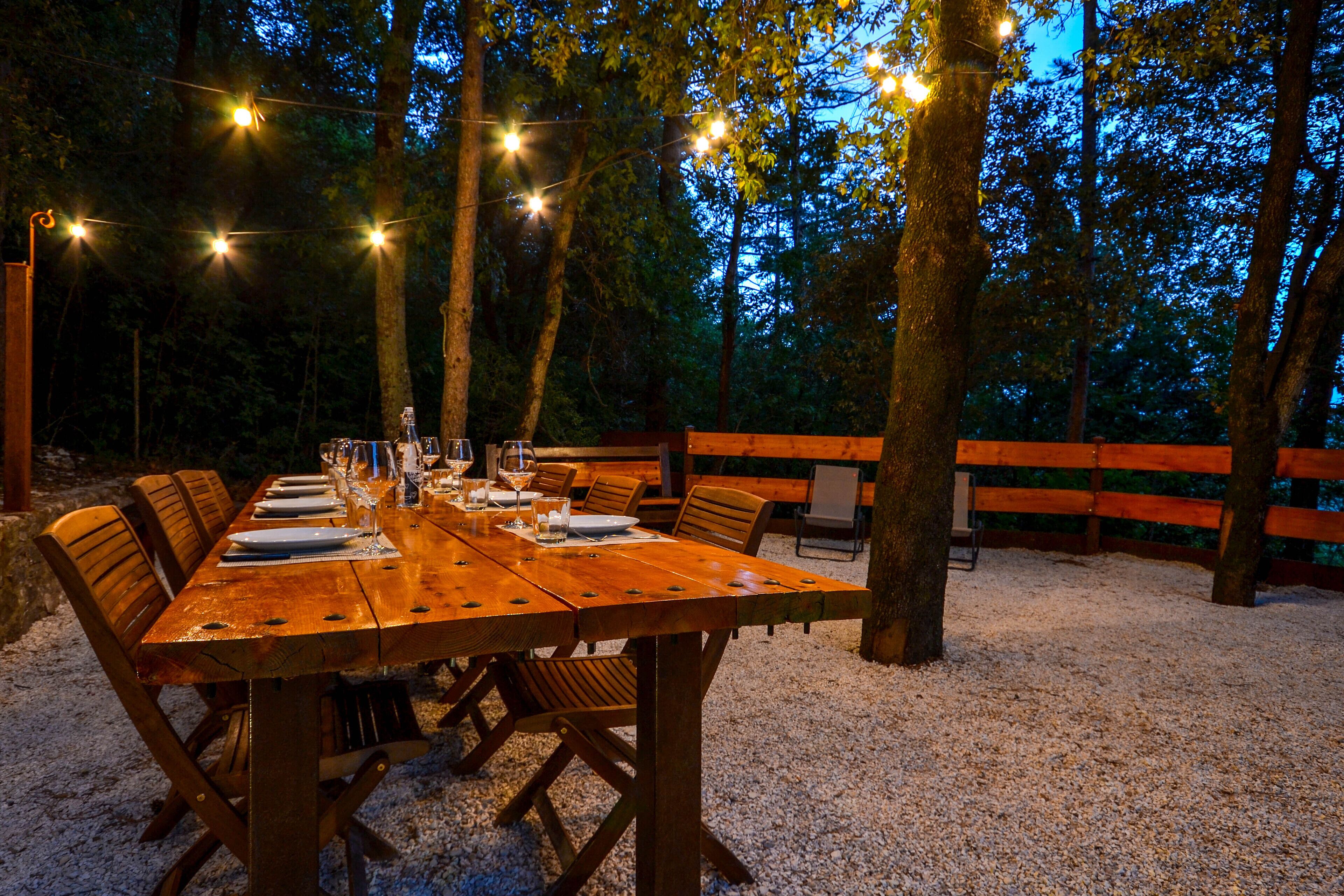 An outdoor wooden table set for dining surrounded by chairs, with string lights overhead and a wooden fence in the background.