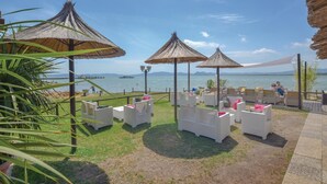 A beachside patio with white wicker furniture and thatched umbrellas, overlooking a calm sea and distant mountains. - Amazing home in Piegaro PG (Piegaro PG)