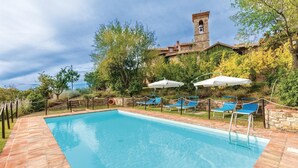 A rectangular swimming pool with clear blue water, surrounded by a stone deck. There are several blue lounge chairs with white umbrellas, and a stone wall with a bell tower in the background. - Amazing home in Piegaro PG (Piegaro PG)