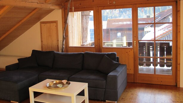 A cozy living room with a dark grey sectional sofa, a white coffee table, and a large window with a view of a wooden house and mountains. - Close to the mountains! (Pfronten)