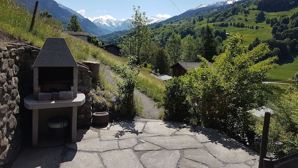 A stone-built outdoor kitchen with a stone bench, surrounded by lush greenery and a mountainous backdrop.