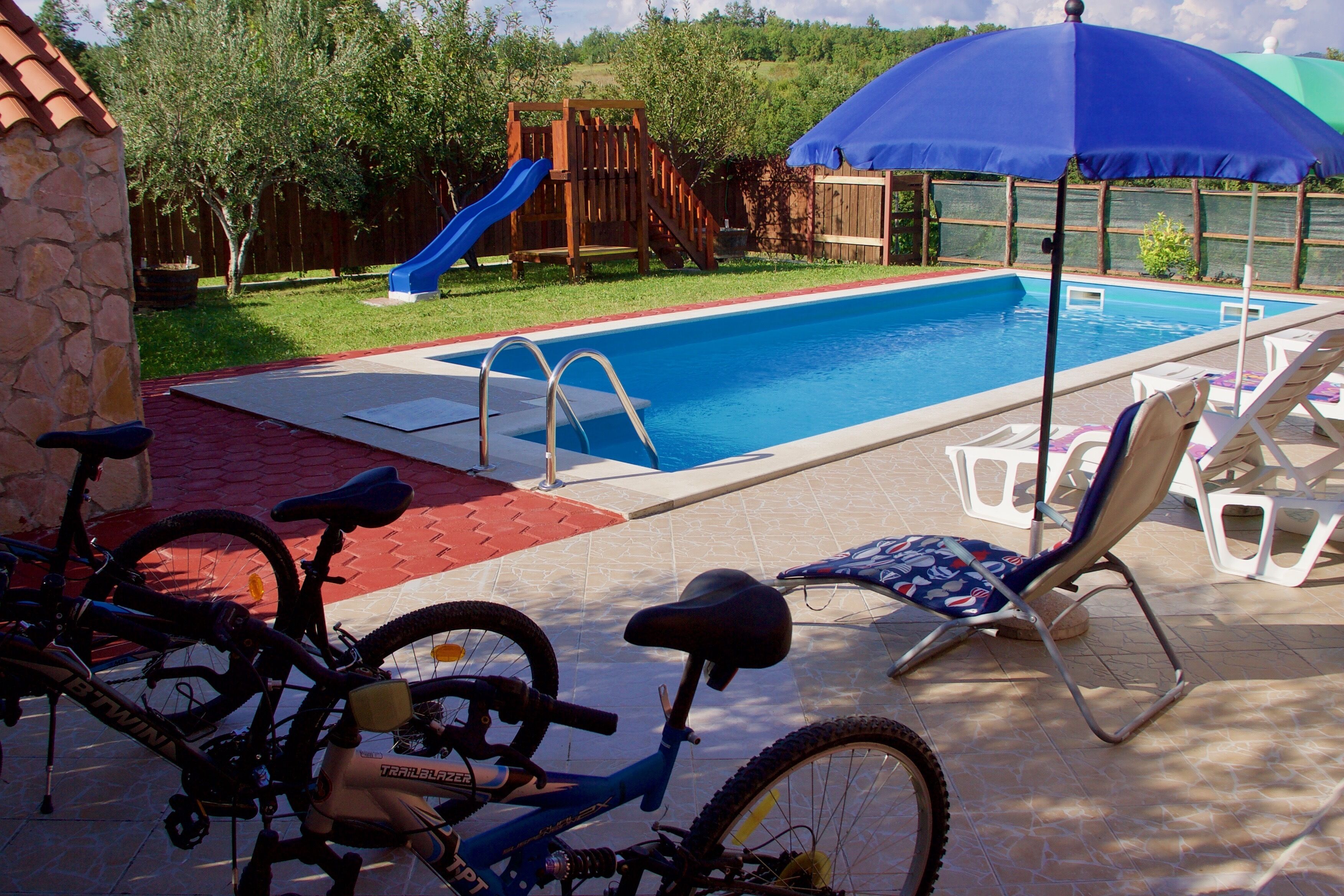 A swimming pool with a slide, surrounded by a patio area with chairs and a bicycle.