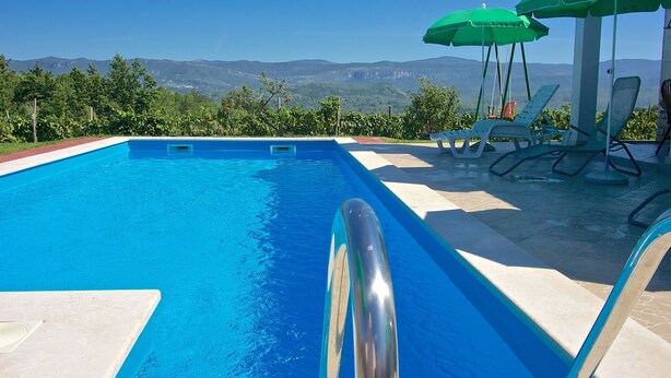Una piscina con agua azul cristalina, rodeada de sillas de descanso y sombrillas verdes, con un paisaje montañoso al fondo.