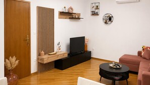 A living room with a wooden floor, a TV on a black stand, a pink sofa, a round black coffee table, and a wooden cabinet with decorative items. - Stunning home in Plat (Plat)