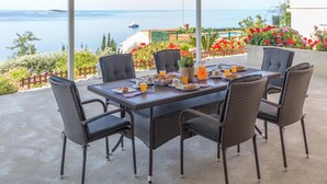 A table set for breakfast with wicker chairs, a view of the sea, and a garden with flowers. - Stunning home in Plat (Plat)