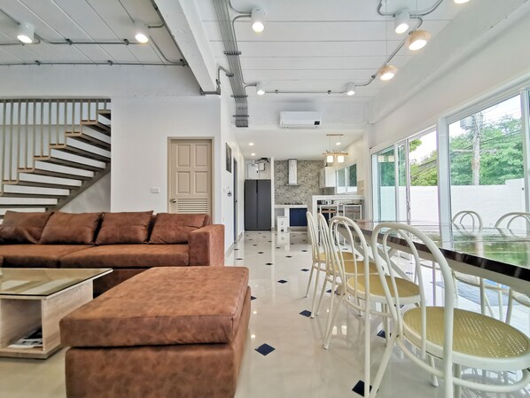 A modern living room with a sofa, ottoman, and dining table with chairs. There is a staircase with a railing, a door, and an air conditioning unit on the wall.