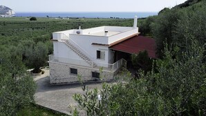 Ein modernes, zweistöckiges Haus mit Natursteinfassade, einem roten Ziegeldach und einem weißen Balkon. Es wird von üppiger Vegetation umgeben und bietet Blick auf eine Küstenlandschaft.