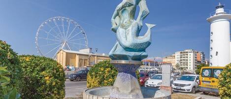 Une statue de sirène dans une fontaine, avec une grande roue et des bâtiments en arrière-plan.