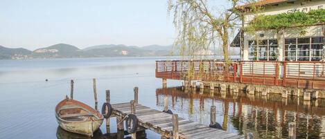 A wooden dock with a boat, a building with a red roof, and a calm lake with mountains in the background.