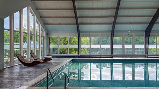Une piscine intérieure avec vue sur la verdure environnante, de grandes fenêtres et des chaises longues.