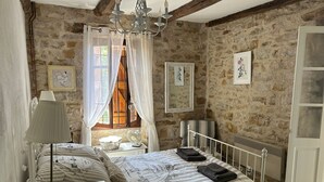 Een slaapkamer met een stenen muur, een bed met gestreept beddengoed, een nachtkastje met een lamp en een raam met gordijnen.