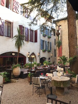 Un espace café en plein air, avec des tables et des chaises, entouré d’immeubles aux volets rouges et bleus, et agrémenté d’un palmier.