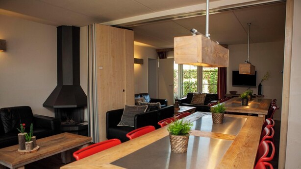 Een moderne woonkamer met een open haard, een eethoek met houten tafels en rode stoelen, en een aan de muur bevestigde televisie.