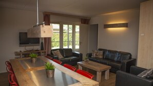 Een moderne woonkamer met een houten eettafel, rode stoelen, een zwarte leren bank en een televisie aan de muur.